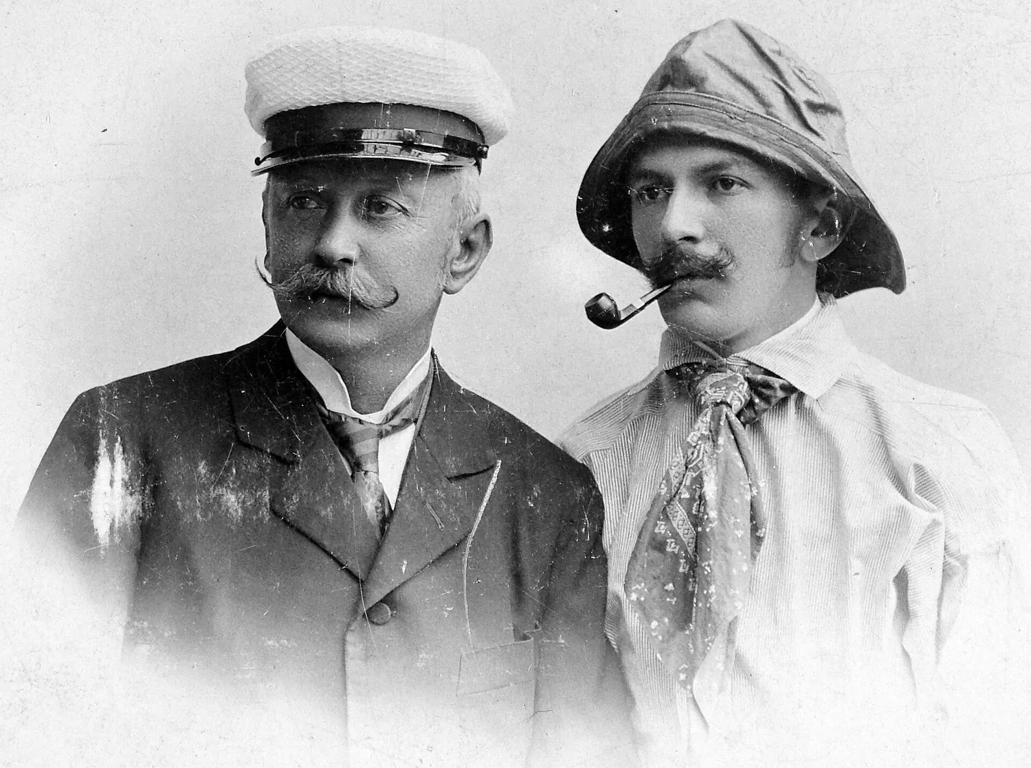 Studio portrait of two men, one in a peaked cap and jacket, the other in a sou'wester with a pipe